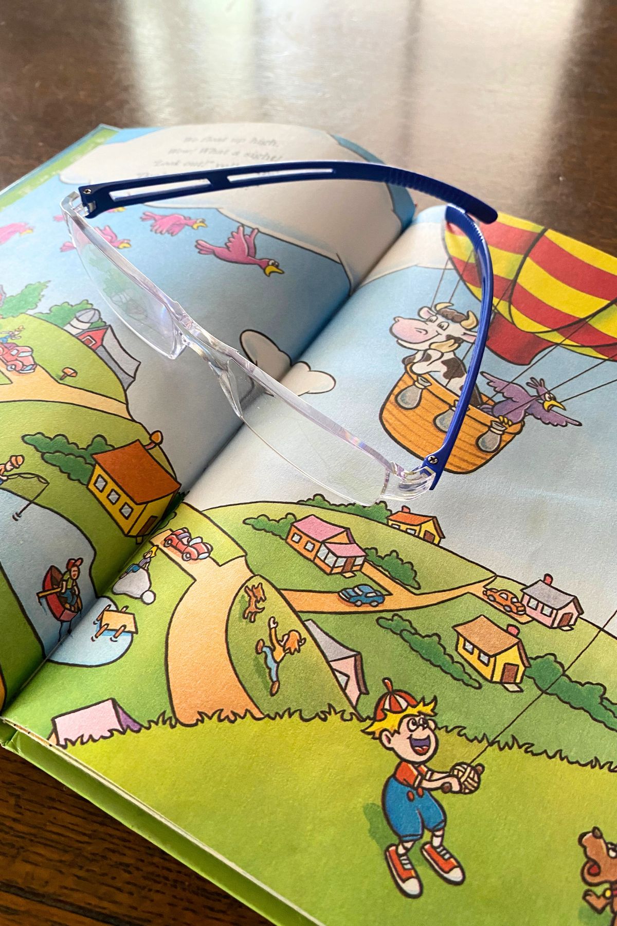 Child's book open to a cartoon cow and bird floating along in a yellow and red striped hot air balloon. A pair of reading glasses is sitting on the book.