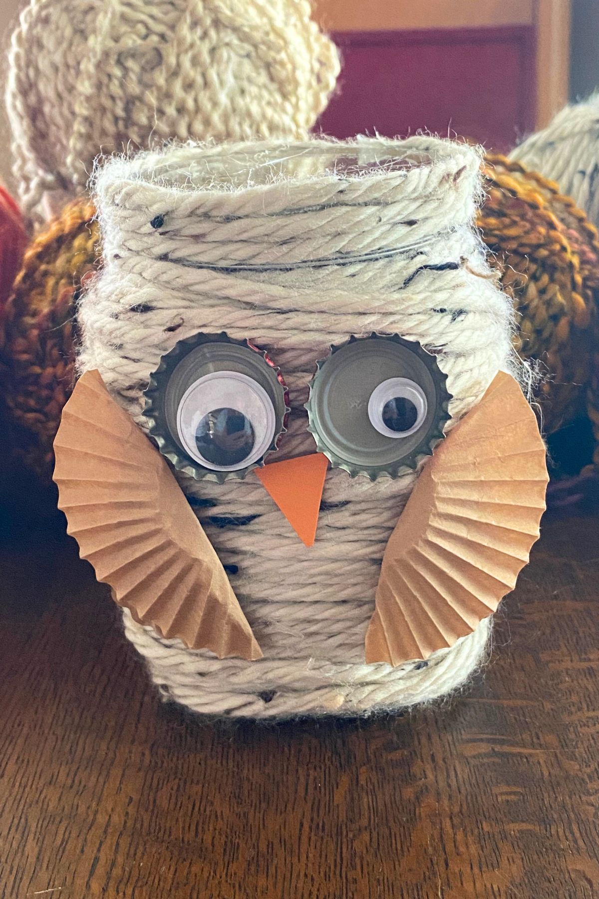 A mason jar wrapped in gray yarn with cupcake wrappers for wings, an orange paper triangle for a beak, bottle-cap google eyes sitting on a wooden table in front of yarn