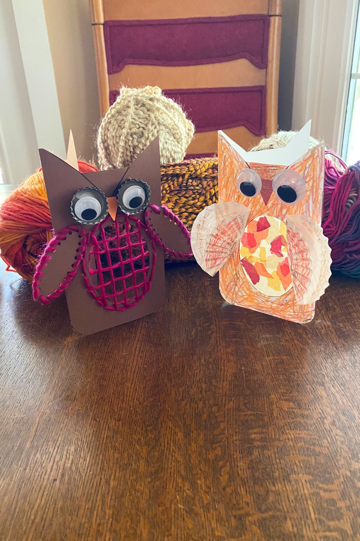 Printable card stock owl crafts. One on brown card stock with pink yarn stitched around the wings and tummy and one with cupcake wrappers for wings and a body colored with crayon sitting on a wooden table in front of yarn