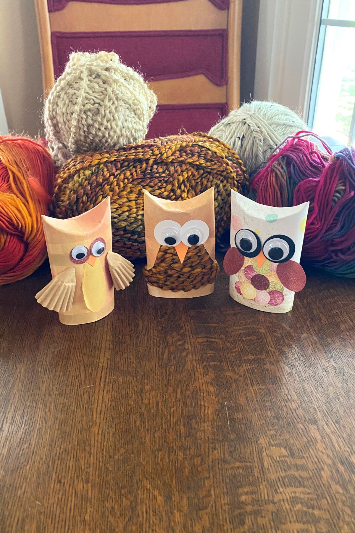 Three paper roll craft owls. One with brown yarn for the body, one with cupcake wrappers for wings and the third with construction paper wings and body sitting on a wooden table with yarn in the background