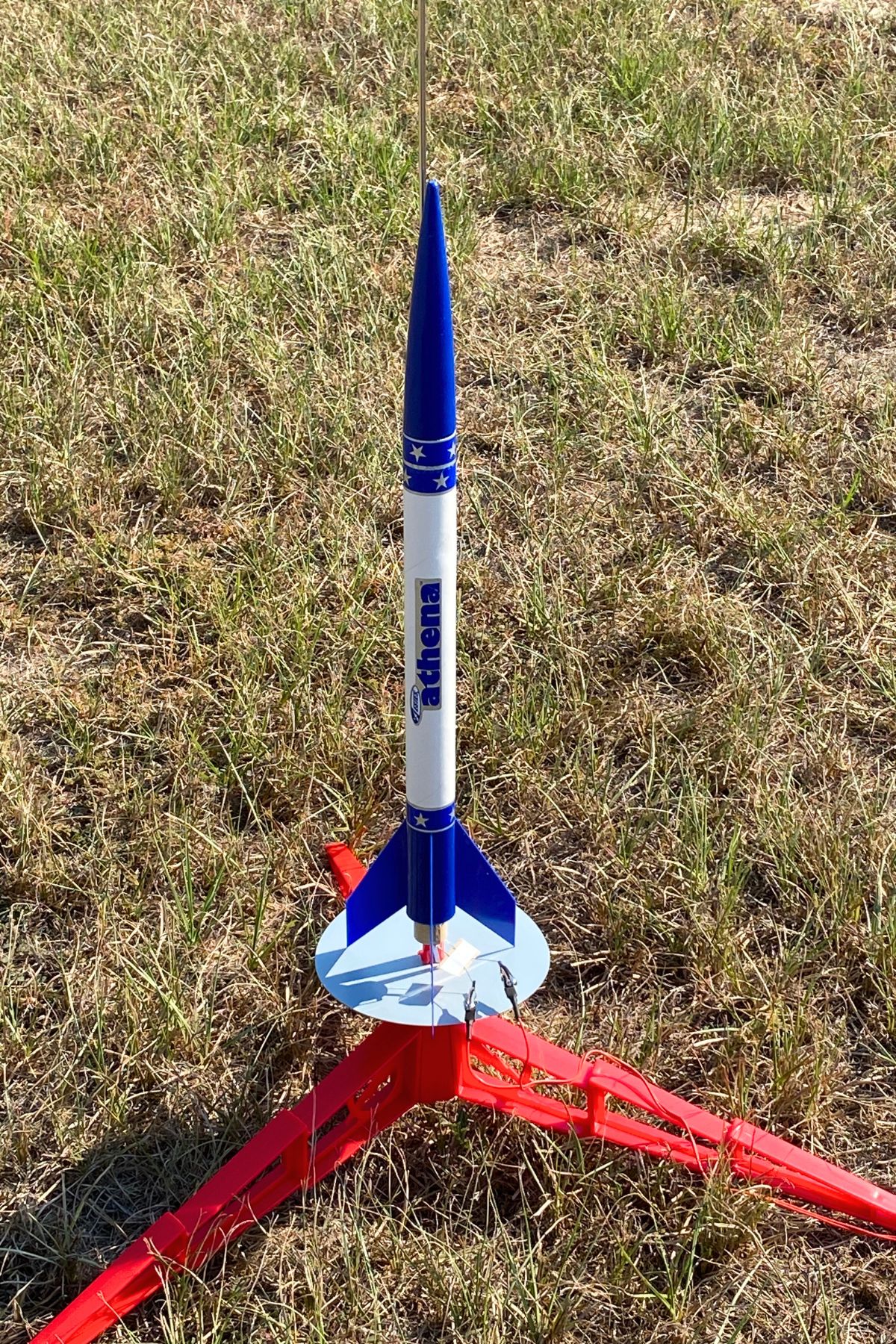 Solid fuel model rocket with white body, blue nose cone and blue fins on a red launch stand
