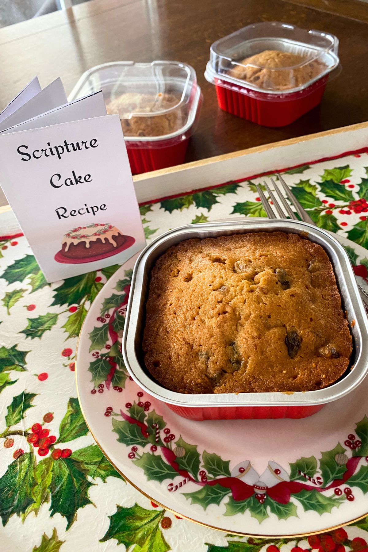 Brown cake with raisins in a square red foil tin on a Christmas plate and fork with two more tins behind on a wooden table