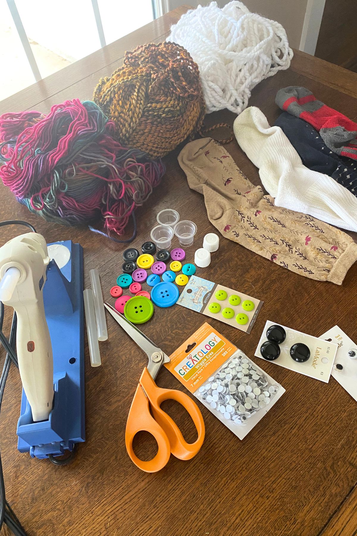 Hot glue gun, thread, yarn buttons, communion cups, oil bottle lids and three socks on a wooden table