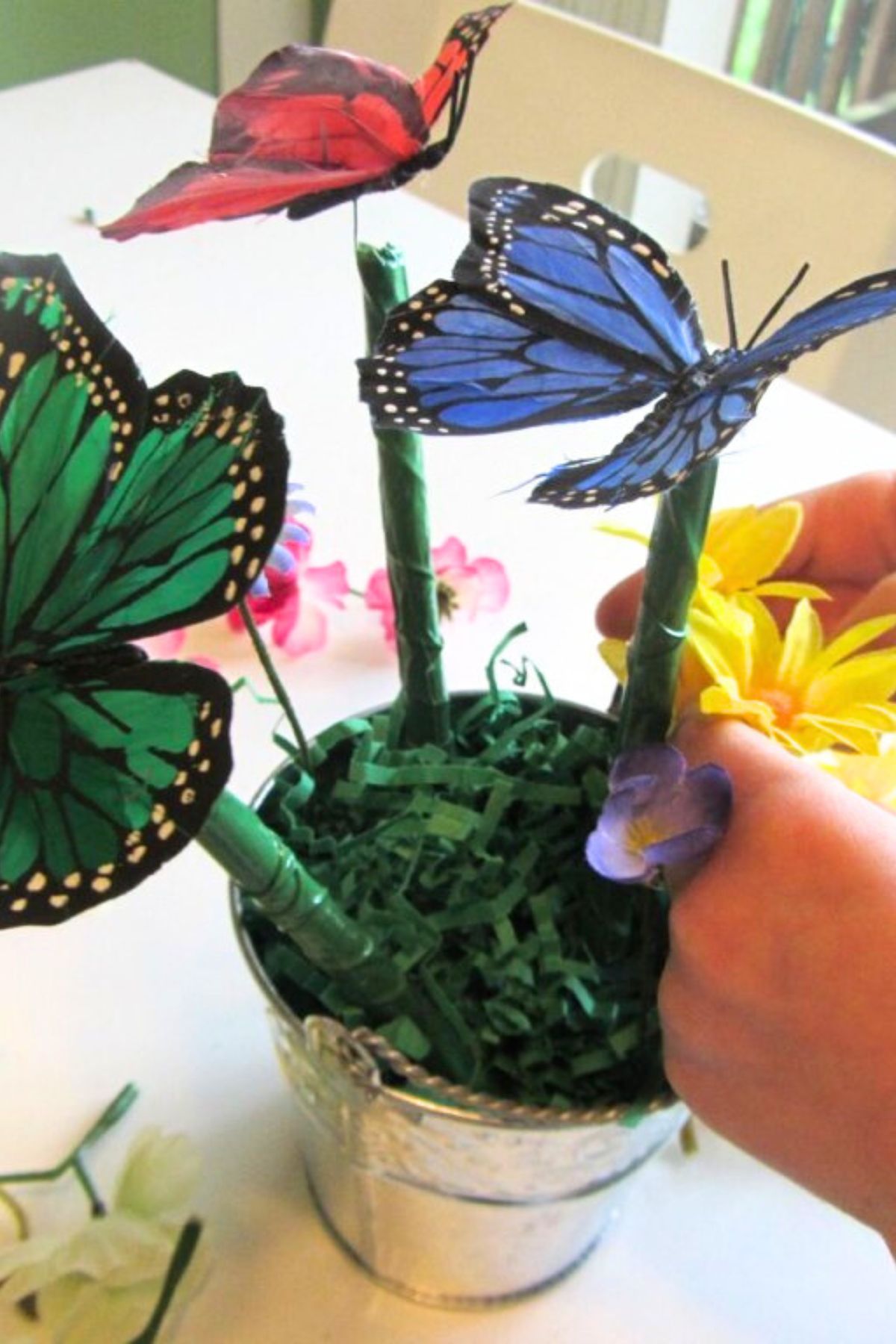 Green, blue and red silk butterflies in a silver bucket with green crinkle grass and colorful silk flowers