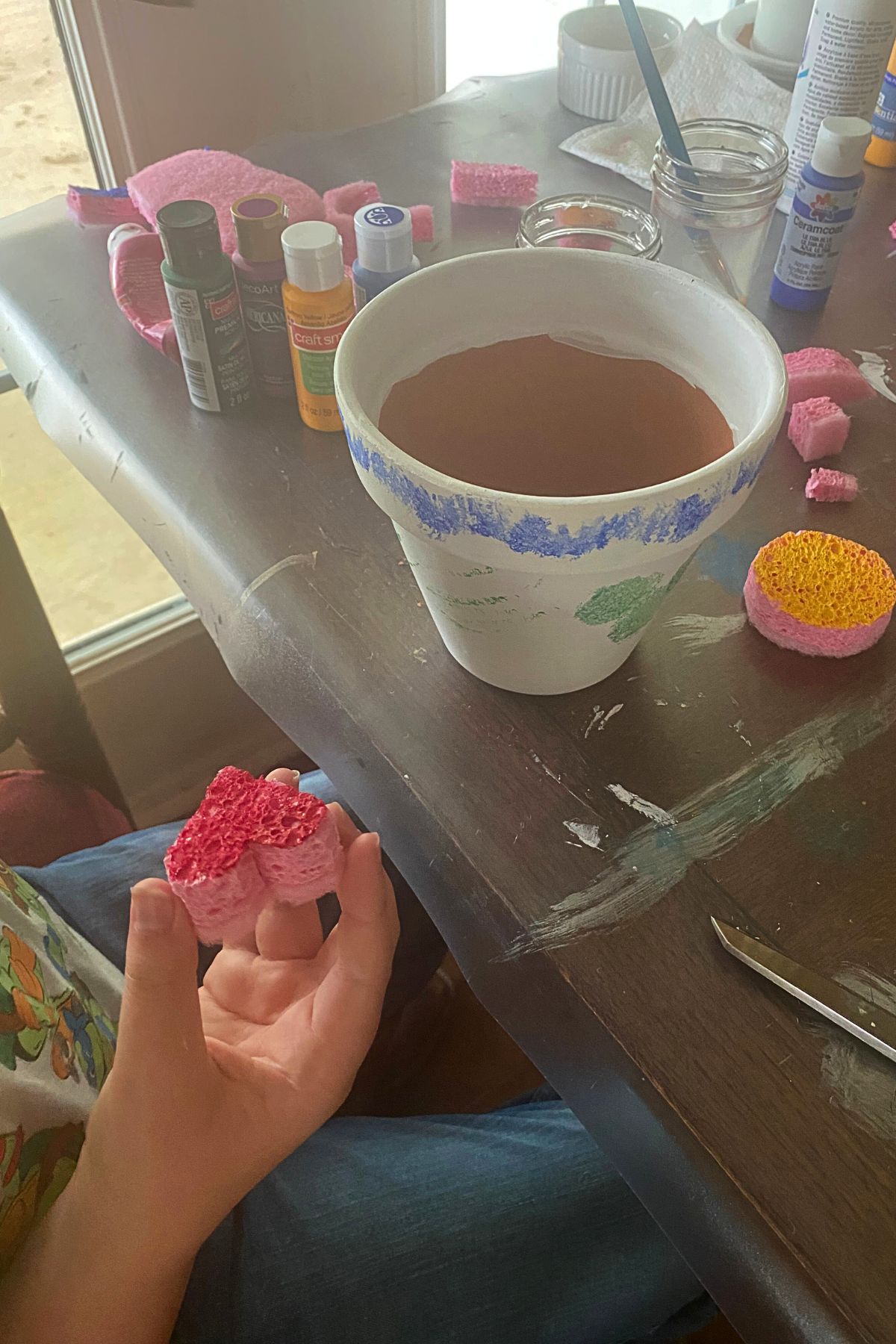 White flower pot on a wooden table with a child's hand brushing paint on a heart-shaped sponge stamp