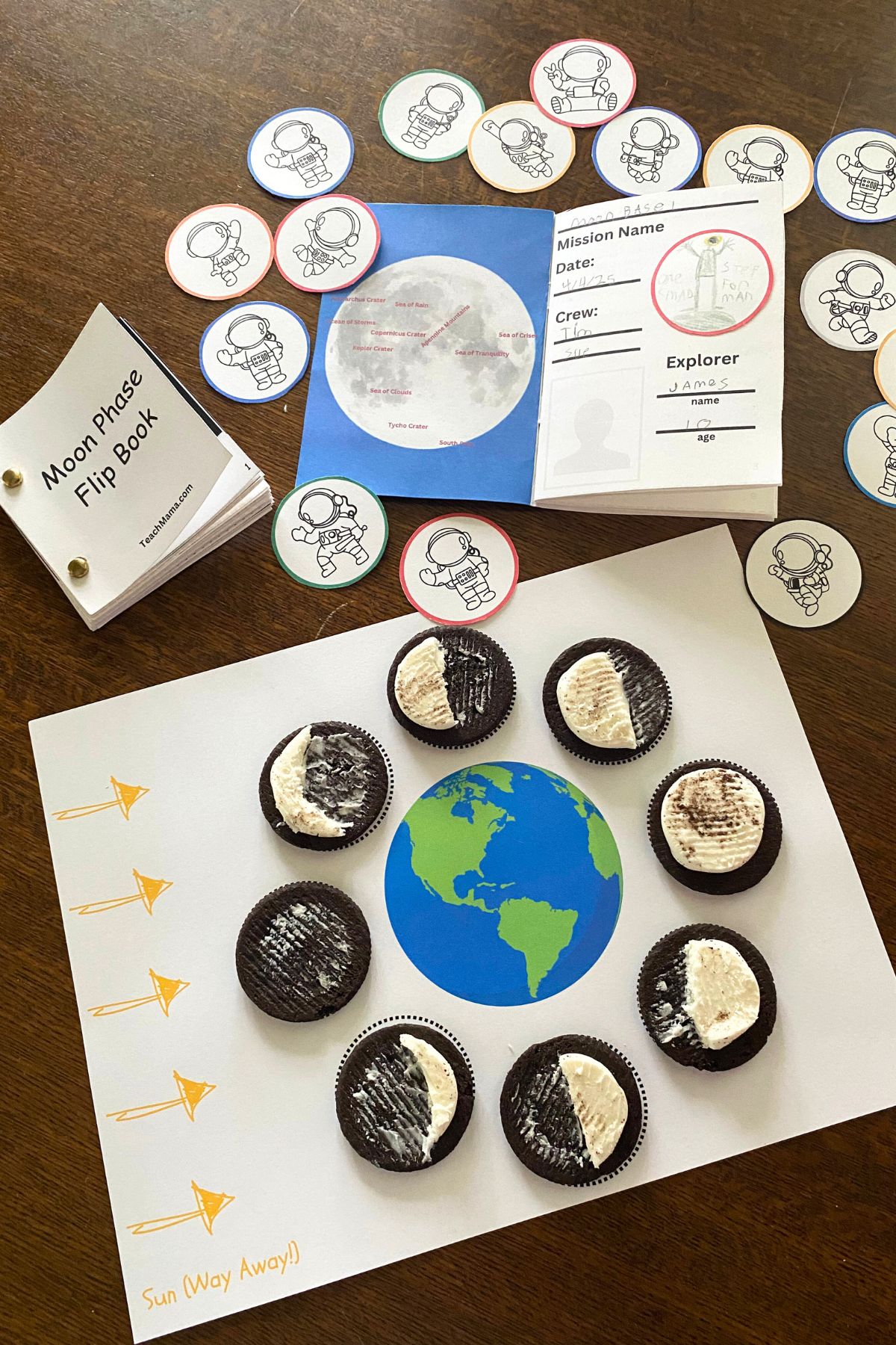 Printed Lunar Passport open to the first page, mission "patches" with cartoon style astronauts, an assembled moon phase flip book and sandwich cookies with the icing carved into moon phases arranged around a printed Earth image