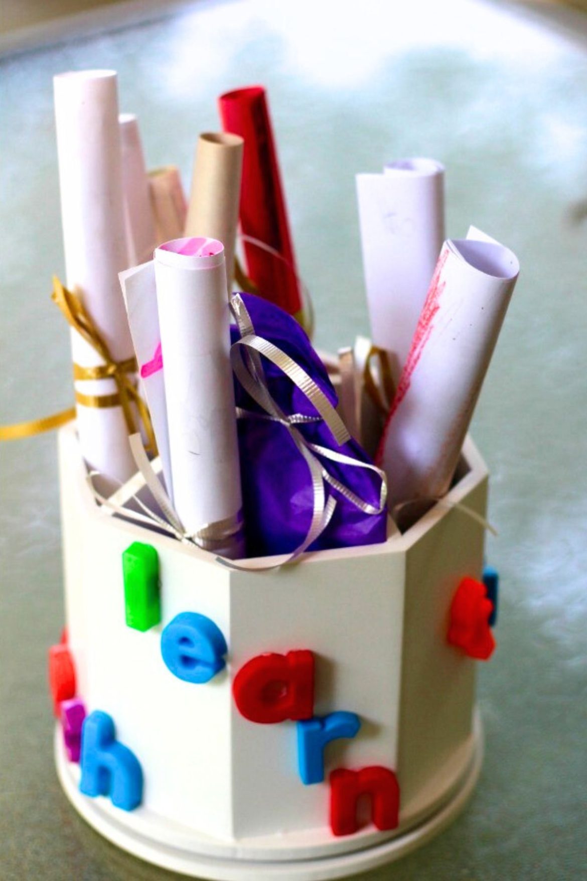 Wooden pencil holder painted white with colorful letters glued to the side and rolled papers and pencils inside