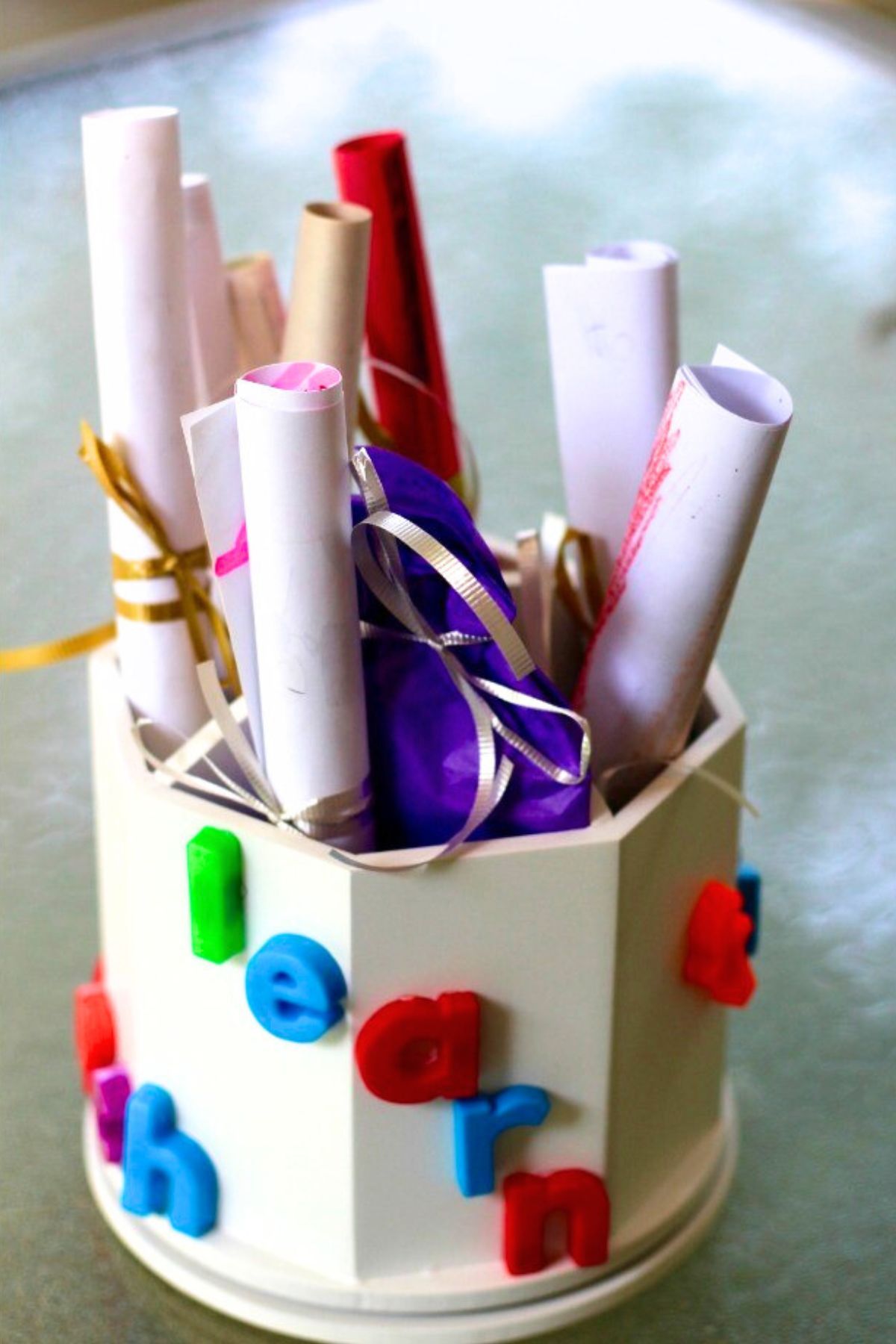 Wooden pencil holder painted white with colorful letters glued to the side and rolled papers and pencils inside