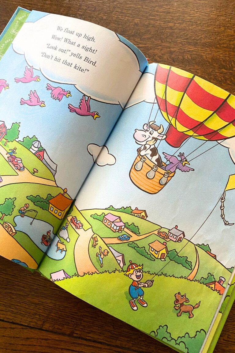 Child's book open to a cartoon cow and bird floating along in a yellow and red striped hot air balloon.