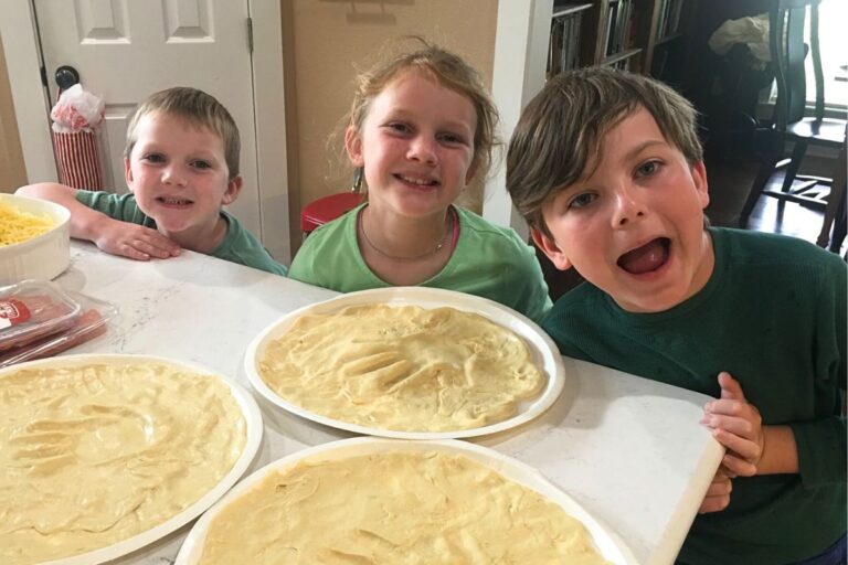 Two young boys and a girl smiling and showing off homemade pizza crust with their handprints in the center