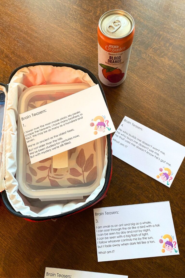 Red soft sided lunch pail with an orange drink and brain teaser lunchbox notes on a wooden table