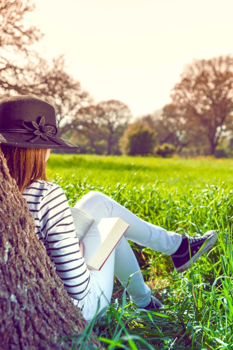 A girl wearing a hat and reading a book under a tree in a grassy field